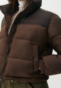 Brun, vadderad jacka med hög krage, med en mörkbrun överdel. Innehåller dragkedja framtill, ribbade mudd på ärmarna och en broderad logga på bröstet.