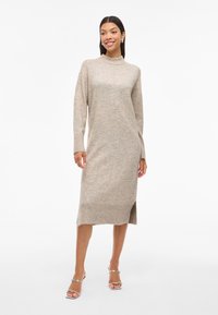 Világos bézs kötött ruha tölcsérnyakúval, hosszú ujjal, oldalsó hasítékkal, bordázott mandzsettával és szegéllyel. Simább tapintású, visszafogott dizájn.