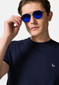 Słoneczne okulary w niebieskim odcieniu z metalową ramką, odbijające światło. Noszone przez osobę w granatowej koszulce z małym białym logo na piersi.