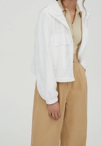 Žena v bílé bundě s kapucí, béžovém žebrovaném zipovém topu a volných béžových kalhotách s vysokým pasem na jednobarevném pozadí.