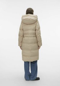 Manteau matelassé beige avec une taille cintrée et une grande capuche. Extérieur lisse avec un motif matelassé, associé à un jean à jambes larges et des chaussures noires.