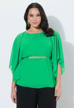 Kvinde med mørkt hår iført en lysegrøn bluse med flagrende ærmer, guldbælte og lange guldringe, stående foran en hvid baggrund.