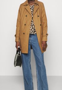 Osoba v béžovém dvouřadém trenčkotu, leopardím topu, modrých džínách a držící černou kabelku.