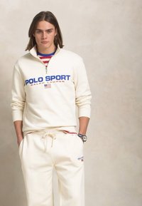 Hanorac crem cu guler cu fermoar, având logo-ul albastru "POLO SPORT" și accente roșii. Asortat cu pantaloni de trening crem, amândouă fiind dintr-un material neted.