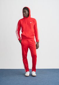 Rdeča jopa s kapuco z belimi črtami na rokavih in logotipom, kombinirana z rdečimi športnimi hlačami. Model nosi bele superge na enostavni podlagi.