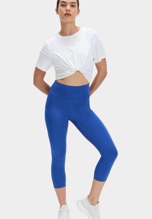 Kobieta stoi z rękami za plecami, ubrana w biały skręcony krótki top, niebieskie legginsy z wysokim stanem i białe trampki na jednolitym tle.