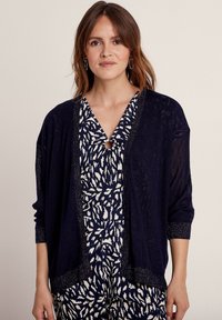 Marineblauwe lichtgewicht cardigan met open voorkant, geribbelde textuur en metallic rand. Gedragen over een gemusterd marineblauw en witte jumpsuit.