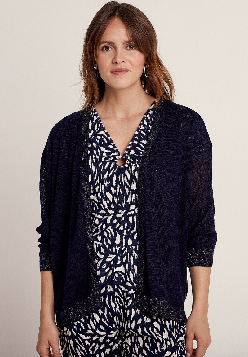 Marineblauwe lichtgewicht cardigan met open voorkant, geribbelde textuur en metallic rand. Gedragen over een gemusterd marineblauw en witte jumpsuit.