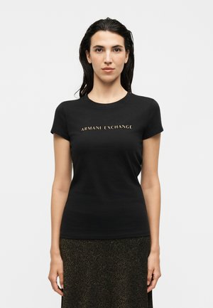 Femme aux cheveux foncés portant un t-shirt noir ajusté avec l'inscription dorée « ARMANI EXCHANGE » et une jupe texturée foncée sur un fond uni.