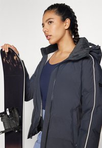 Veste isolante bleu foncé avec une capuche texturée et des finitions blanches contrastantes, portée par une personne tenant un snowboard noir.