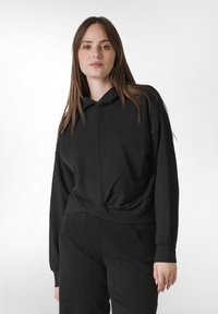 Svart huvtröja i mjukt tyg, med ett framstycke som har detaljer och långa ärmar. Kombinerad med höga svarta byxor med fickor.