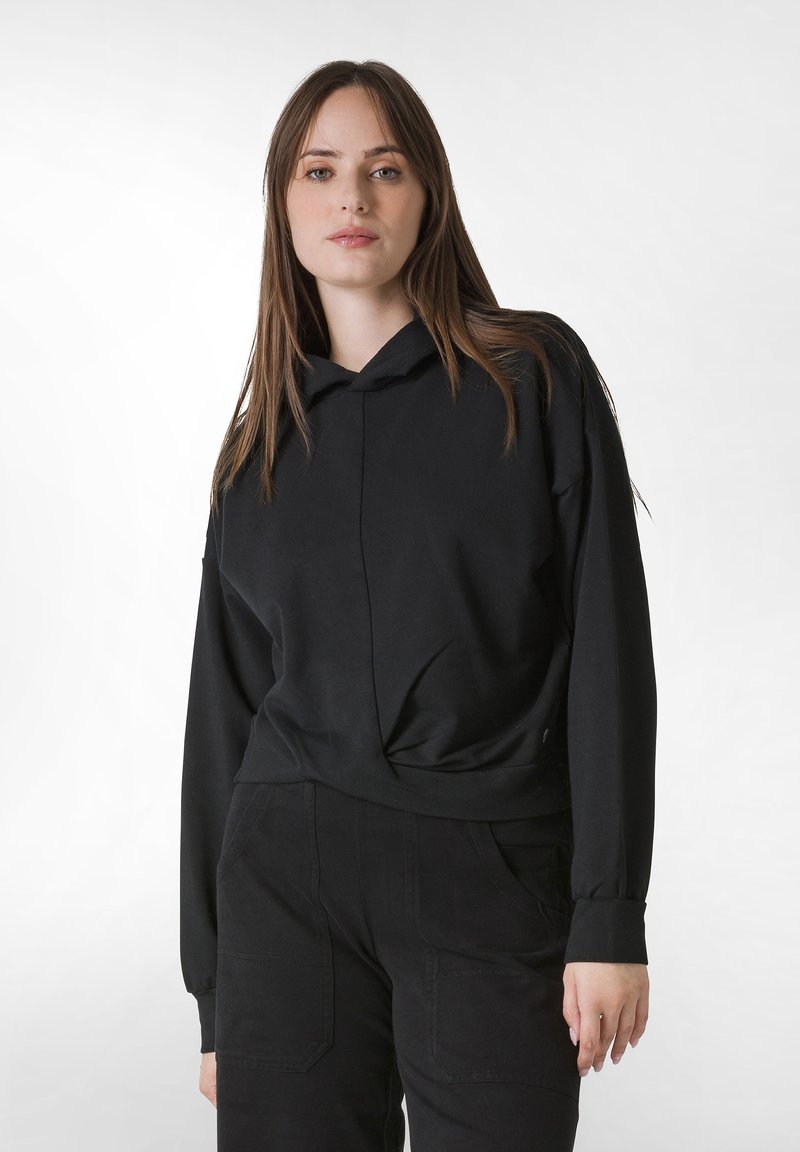 Svart huvtröja i mjukt tyg, med ett framstycke som har detaljer och långa ärmar. Kombinerad med höga svarta byxor med fickor.