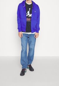 Lila zip-up hoodie över en svart grafisk t-shirt, i kombination med ljusblå jeans och svarta sneakers med rosa och teal-accenter.