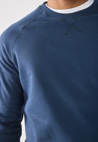 Man in een blauwe crew neck sweatshirt met zichtbare stitchdetails bij de kraag en schouder tegen een effen achtergrond.