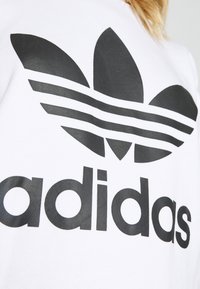 Vit bomulls sweatshirt med en framträdande svart Adidas-logotyp med tre ränder och bladliknande designelement. Slät textur.