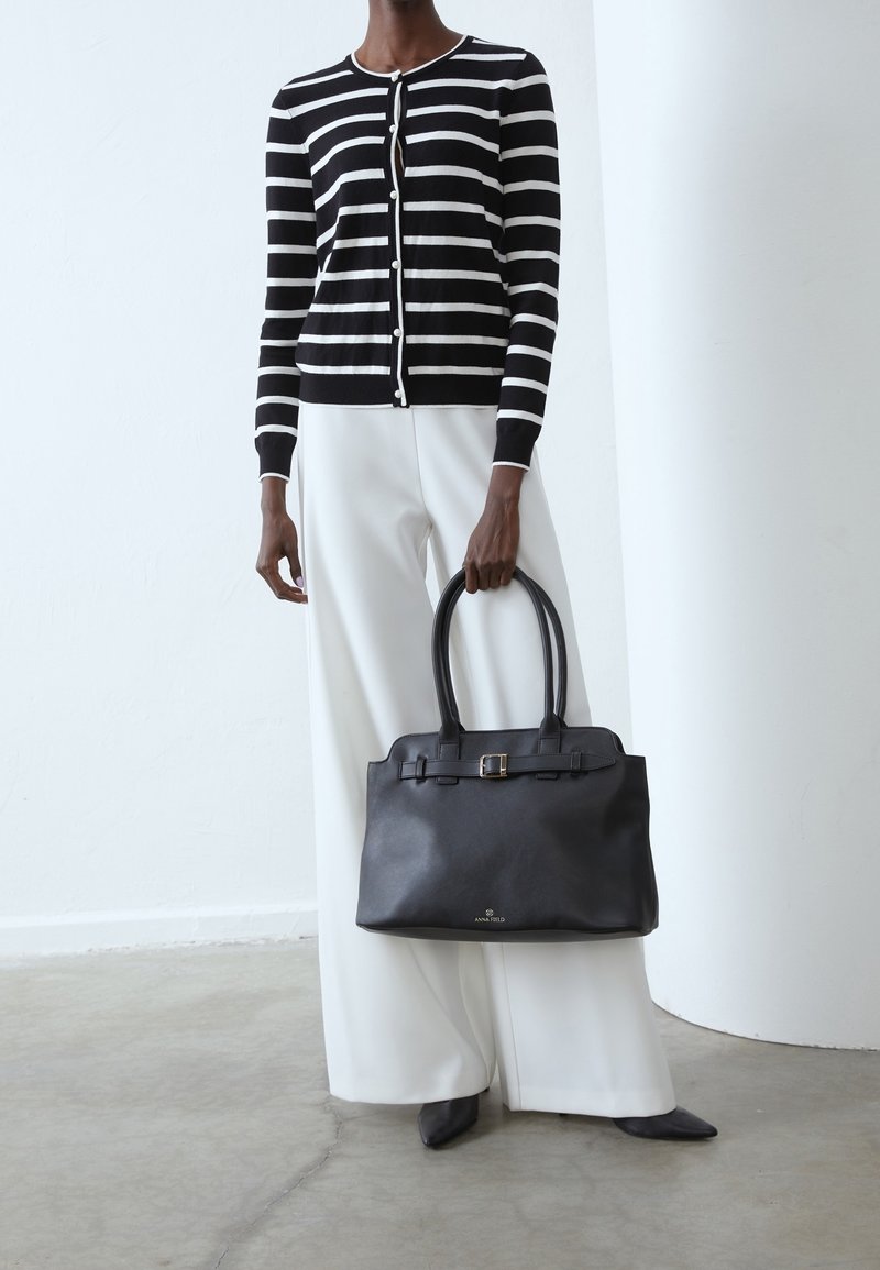 Personne portant un cardigan rayé noir et blanc et un pantalon blanc à jambes larges tient un grand sac à main en cuir noir avec des doubles poignées.