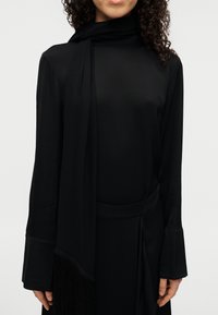 Μαύρο μακρυμάνικο τοπ με ψηλό λαιμό και ένα ριγέ μαντήλι. Μαλακό ύφασμα με διακριτικό γυάλισμα, με διακοσμητικό κρόσσι στο τελείωμα.