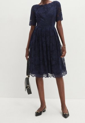 Nainen pukeutuneena polvipituiseen laivastonsiniseen pitsimekkoon, jossa on lyhyet hihat, kädessään musta käsilaukku ja jalassaan mustat matalakorkoiset kärjelliset kengät, joissa on kultainen yksityiskohta.