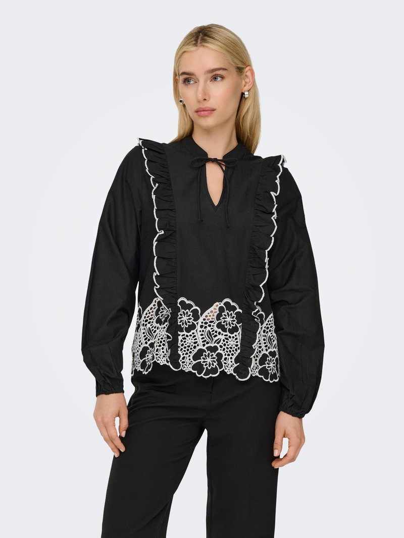 Vrouw draagt een zwarte blouse met lange mouwen met gerimpelde voorpanelen en witte bloemenkantborduursel aan de zoom, gecombineerd met een zwarte broek.