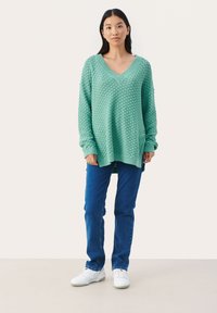 Menta zöld, oversized V-nyakú pulóver texturált mintával. Kék farmer nadrággal és fehér sportcipővel párosítva. Egyszerű, hétköznapi megjelenés.