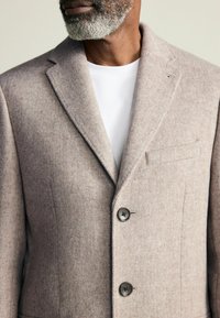 Jasnobrązowa marynarka z mieszanki wełny z klapami, dwoma czarnymi guzikami i kieszenią na piersi. Teksturowana powierzchnia, minimalistyczny design.