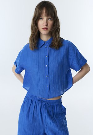 Jeune femme aux cheveux bruns mi-longs portant une chemise ample texturée bleu cobalt boutonnée et un pantalon assorti à taille élastique sur fond blanc.