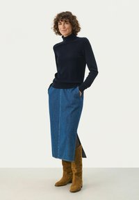 Marineblauer Rollkragenpullover kombiniert mit einem langen Jeansrock mit seitlichem Schlitz und braunen Wildleder-Stiefeln bis zum Knie. Glatte Textur insgesamt.