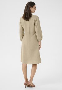 Bežinė lininė suknelė su trijų ketvirčių pūstomis rankovėmis, juosmens siūle ir žemiau kelių ilgiu. Modelis, vilkintis rudus mules, stovi ant šviesaus fono.