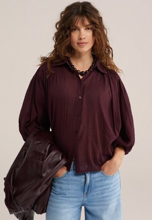 Femme aux cheveux bouclés portant un chemisier bordeaux à boutons, un jean bleu clair, un collier noir à perles, tenant une veste en cuir foncé.