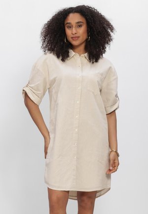 Vrouw met krullend haar die een beige blousejurk met opgerolde mouwen draagt, gouden creooloorbellen en een gouden armband.