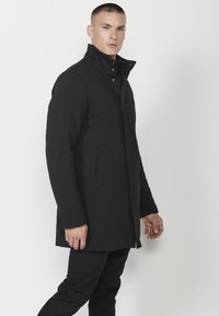 Cappotto in misto lana nero con colletto alto, tasche a pattina e design dritto, dotato di chiusura a bottone e cerniera.