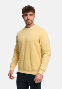 Gul bomullsgenser med rund hals og ribbede mansjetter. Har en liten hvit logo på brystet. Brukes med mørke bukser.