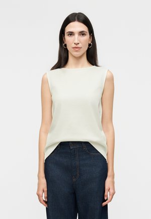 Γυναίκα με ίσια σκούρα μαλλιά φοράει αμάνικη ανοιχτό μπεζ μπλούζα που έχει βάλει μέσα σε σκούρο μπλε ψηλόμεσο τζιν, στέκεται μπροστά σε απλό φόντο.