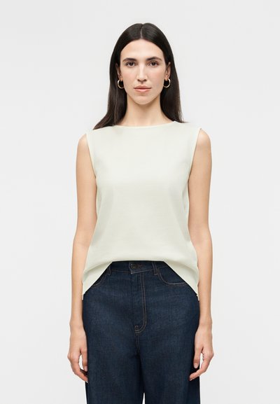 Femme aux cheveux noirs et raides portant un haut sans manches beige clair rentré dans un jean taille haute bleu foncé, debout devant un fond uni.