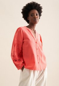 Koronowy koszula zapinana na guziki z dekoltem w kształcie litery V, ozdobiona haftowanymi kwiatowymi wzorami na bufiaste rękawach oraz o luźnym kroju. Materiał wydaje się lekki.