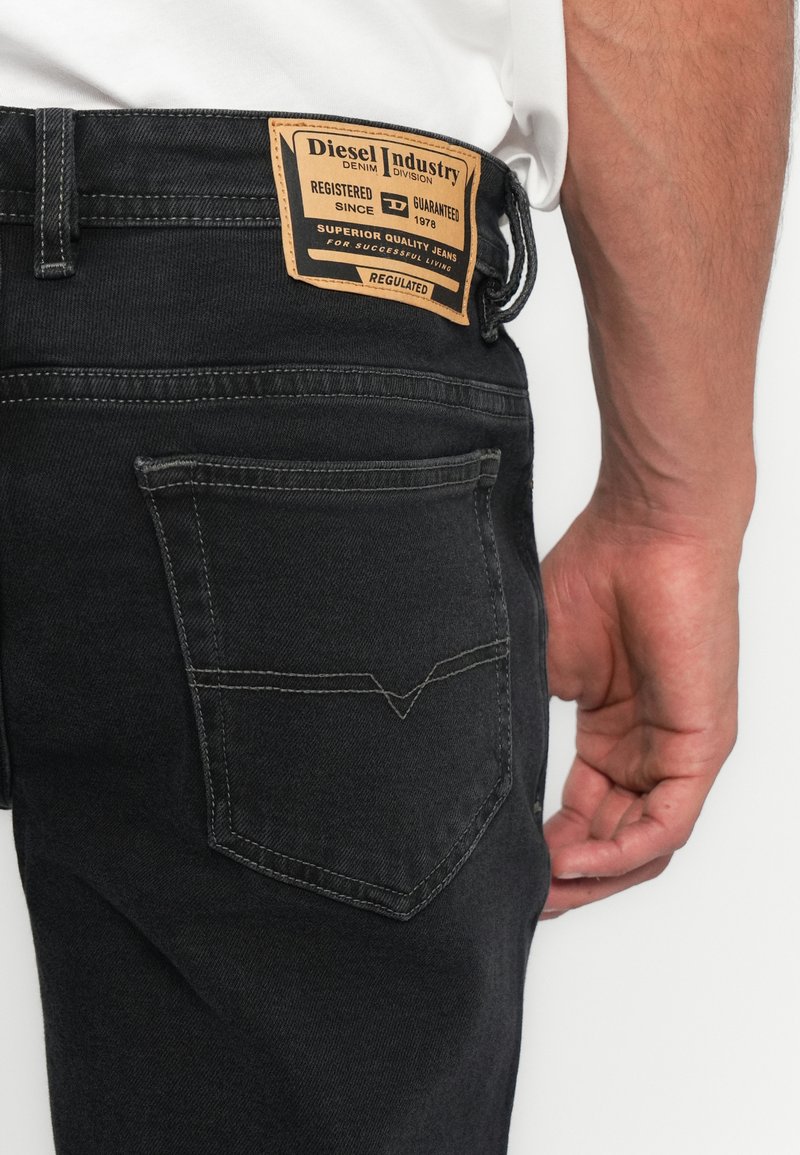 Schwarz Jeans mit Leder-Logo-Patch in der Gesäßtasche, getragen von einer Person in einem weißen Hemd, deren Hand entspannt an der Seite hängt.