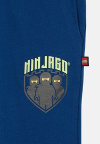 Blue cotton sweatpants featuring a Ninjago logo and graphic of three masked figures in gray, with a small red LEGO tag on the side.