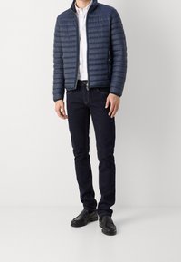 Giubbotto trapuntato blu navy con colletto alto, abbinato a una camicia bianca e jeans scuri. Mocassini neri completano il look. Tasche con cerniera semplici.