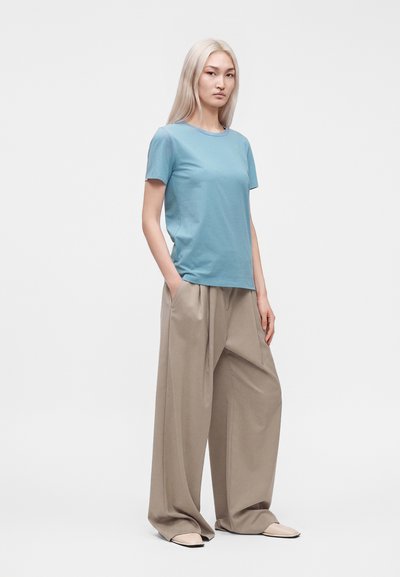 Femme aux longs cheveux blonds portant une chemise bleue à manches courtes et un pantalon beige large, debout devant un fond uni.