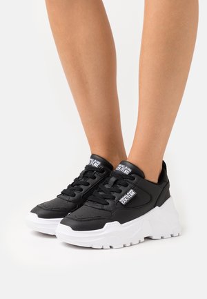 Baskets noires Versace Jeans Couture avec semelles blanches épaisses portées sur des jambes nues sur fond blanc.