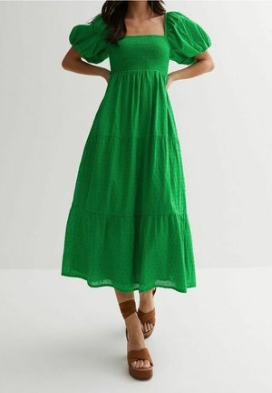 Femme debout, mains sur les hanches, portant une robe midi verte à manches bouffantes et des sandales compensées marron sur fond blanc uni.