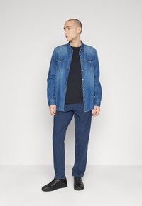 Camicia in denim con lavaggio blu scuro, bottoni a pressione e due tasche sul petto, indossata sopra una maglietta nera, abbinata a pantaloni navy e sneakers nere.