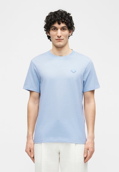 Mladi moški s kodrastimi temnimi lasmi, oblečen v svetlo modro majico s kratkimi rokavi z majhnim emblemom srca na prsih, stoji ob enobarvnem ozadju.