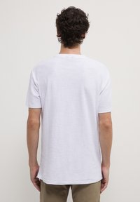 Uomo con capelli castani ricci che indossa una semplice camicia bianca a maniche corte e pantaloni beige, visto di spalle su uno sfondo semplice.