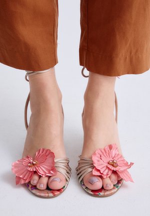 Pies que llevan sandalias de punta abierta con grandes decoraciones de flores rosadas y tiras beige delgadas, combinados con pantalones color óxido.