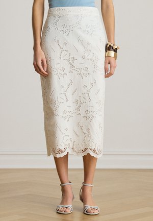 Femme portant une jupe midi en dentelle blanche, un haut bleu clair, des talons beiges à lanières et un gros bracelet au poignet droit.