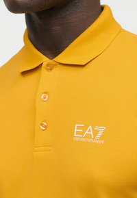 Col avec col et boutons d'un polo jaune moutarde avec logo blanc "EA7 Emporio Armani" sur la poitrine, porté par une personne à la peau foncée.