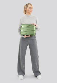 Vrouw in een grijze trui en broek die een kleine groene koffer vasthoudt tegen een effen lichte achtergrond, kijkend naar de zijkant.