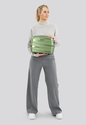 Vrouw in een grijze trui en broek die een kleine groene koffer vasthoudt tegen een effen lichte achtergrond, kijkend naar de zijkant.