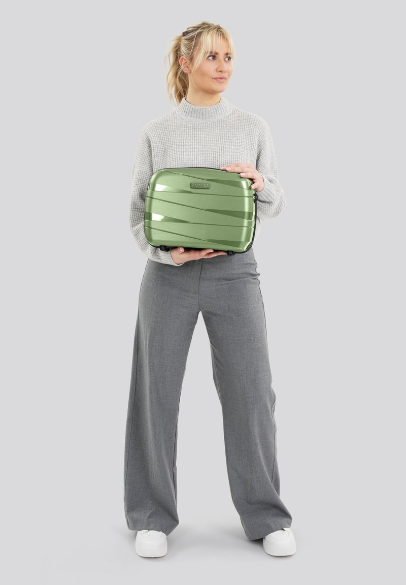 Vrouw in een grijze trui en broek die een kleine groene koffer vasthoudt tegen een effen lichte achtergrond, kijkend naar de zijkant.